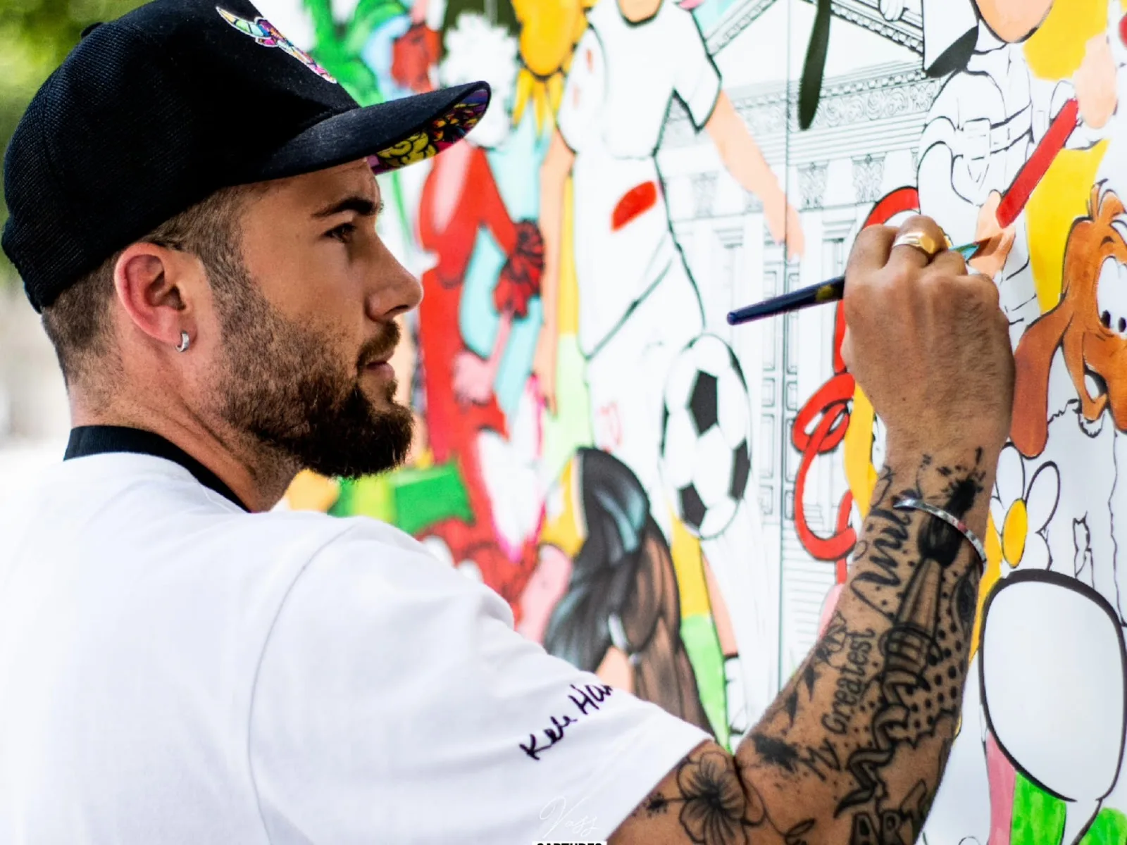 Melvyn Barros en train de peindre une grande fresque colorée, casquette noire et bras tatoué